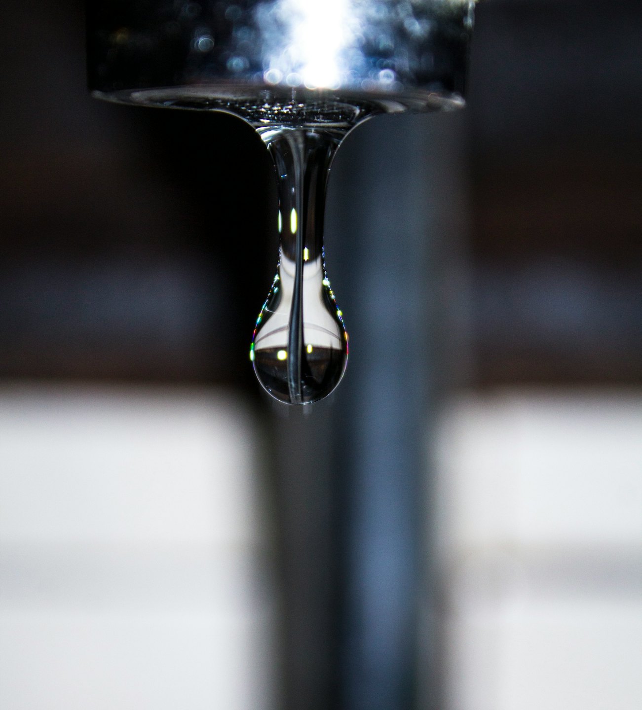 A clear water droplet hangs from a chrome kitchen faucet.